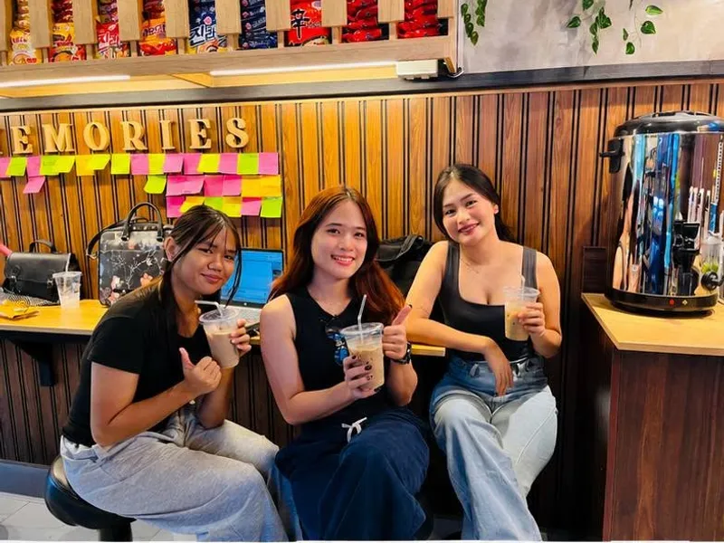 Three friends with iced drinks in front of the MAKE MEMORIES wall at K-Stop